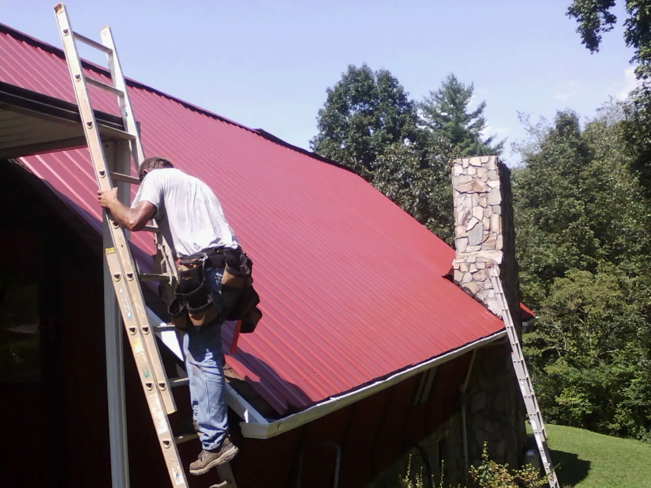 Roofer on a ladder beside a red metal roof and stone chimney — Soffit & Fascia Repair by Roofing Brothers in Santa Monica
